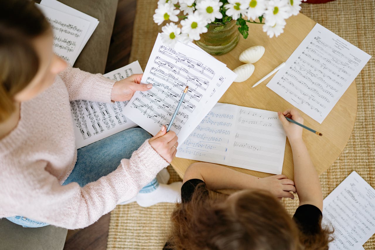Two people studying music with sheet music and notebooks on a table indoors.