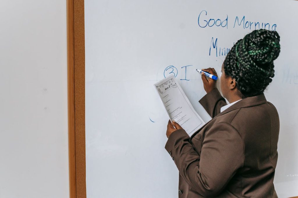A female teacher in a brown suit writes on a whiteboard in a classroom setting.