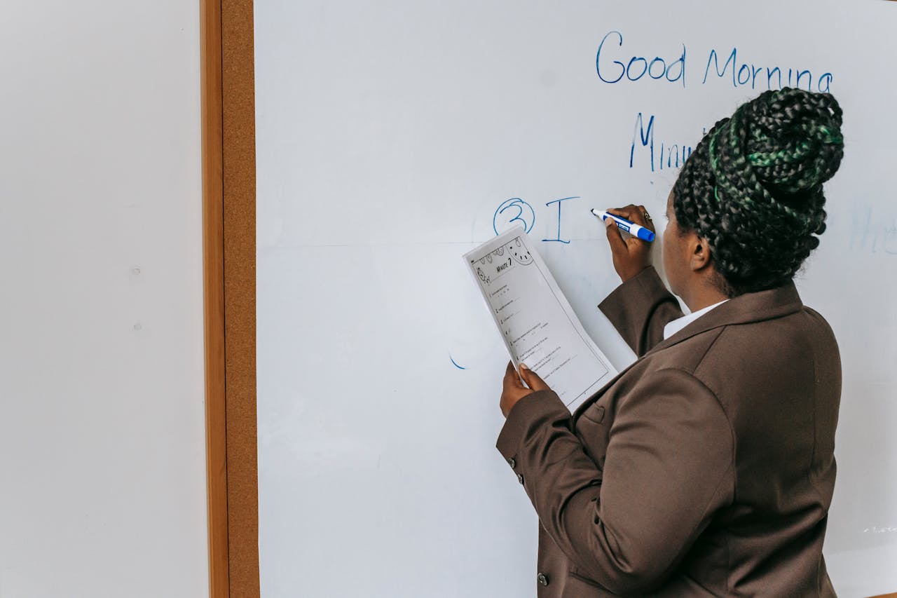A female teacher in a brown suit writes on a whiteboard in a classroom setting.