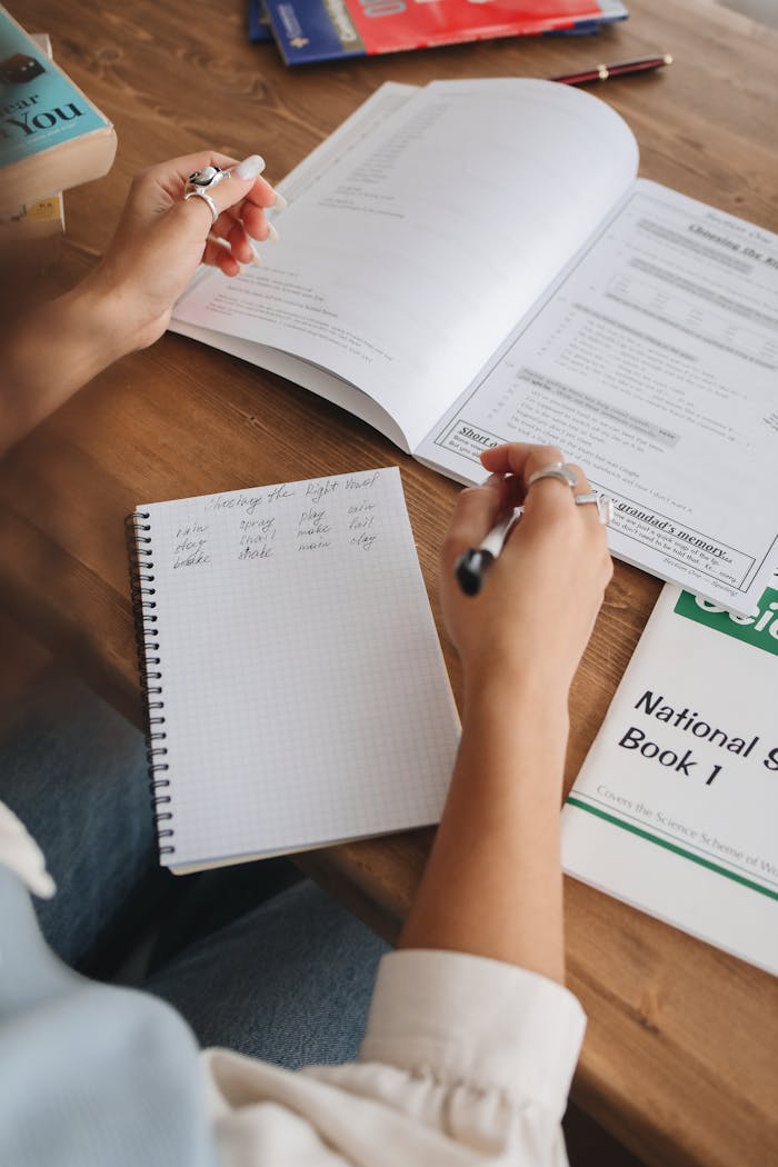 Close-up of a student taking notes while studying with textbooks and notebook on a wooden desk.